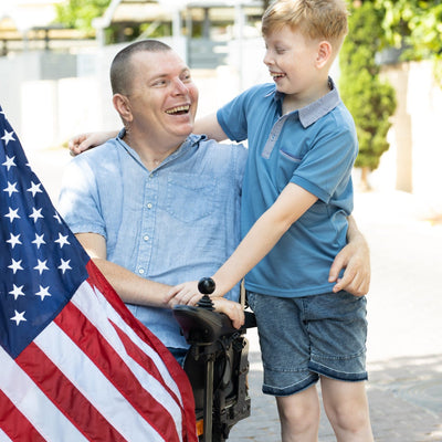 Man in a wheelchair with a young boy holding an American flag, outdoors.